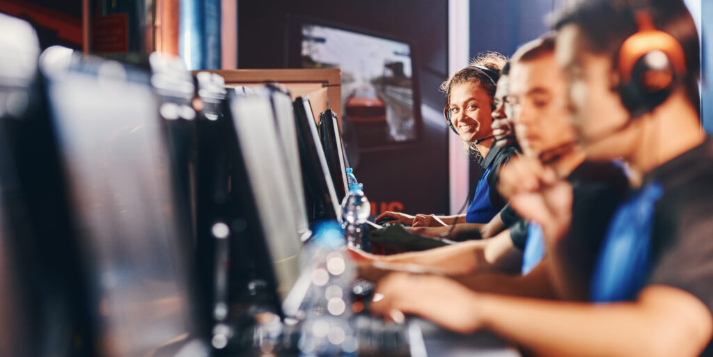Video games online competition. Team of young professional cybersport gamers wearing headphones participating in eSport tournament, sitting in a row in internet cafe. Mixed race girl smiling at camera. (Foto: banco de imagens)